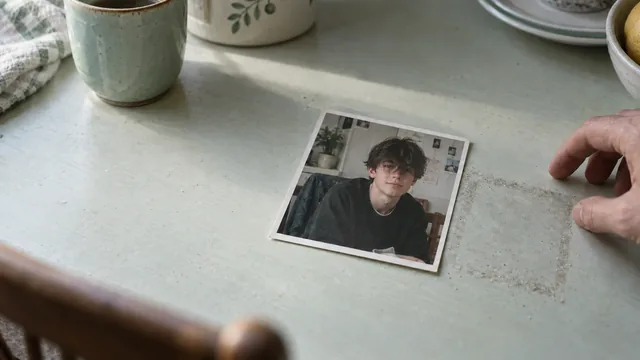A photograph of a boy on a kitchen table, beside a hand and everyday objects.